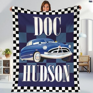 Può includere: Una coperta blu con un'immagine a cartoni animati di Doc Hudson, un'auto blu, con la scritta "DOC HUDSON" in bianco. La coperta ha un bordo a scacchi in bianco e nero.