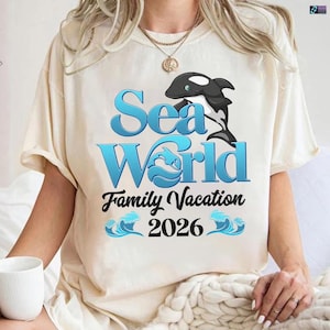 May include: Cream-colored t-shirt featuring the words "Sea World" in blue, with a black and white orca graphic. Below the text is "Family Vacation 2026" in black, with wave graphics. The shirt is a casual style.