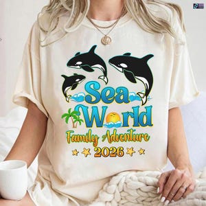 May include: Cream-colored t-shirt featuring a graphic of three black and white orcas, the words "Sea World Family Adventure 2026", a palm tree, a sun, and waves. The design is in shades of blue, yellow, and green.