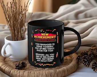May include: A black ceramic mug featuring the text: NEW ACHIEVEMENT! HERE WE FUCKING GO AGAIN! YOU MANAGED TO DRAG YOURSELF OUT OF BED TODAY. GOOD FOR YOU! REWARD: ALL YOU GET IS THIS STUPID MUG. ISN'T BEING AN ADULT FUN? The mug has a handle and colourful accents.