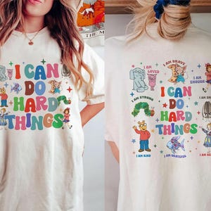 May include: Cream-colored t-shirt with the text "I CAN DO HARD THINGS" in colorful letters. The shirt features cartoon illustrations of various characters and the phrases "I AM BRAVE", "I AM STRONG", and "I AM KIND".