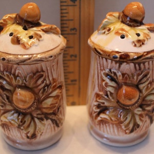 Może przedstawiać: Para solniczek i pieprzniczek ceramicznych w kształcie pni drzew z wierzchołkami w kształcie żołędzi. Solniczki są ozdobione brązową i białą glazurą i mają kwiatowy wzór.