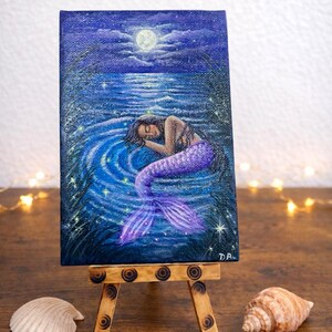 May include: A small canvas painting on a wooden easel depicts a mermaid sleeping in blue water under a full moon. The mermaid has a purple tail and dark hair. Seashells and crystals are arranged around the easel.