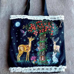 May include: A black canvas tote bag with a white lace trim. The bag features a hand-painted design of a tree with red apples, a deer, a rabbit, a hummingbird, and a squirrel. The background is a dark blue with white stars.