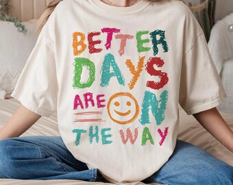 May include: A white t-shirt with the text "Better Days Are On The Way" in colorful, hand-drawn lettering. A smiley face is included in the design.