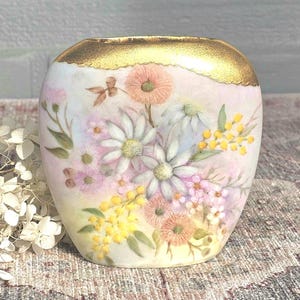 May include: A small, hand-painted vase with a gold rim and a floral design. The vase features a variety of flowers in shades of pink, white, yellow, and peach. The vase is approximately 10 cm tall and 10 cm wide.
