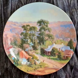 May include: A decorative plate with a landscape painting of a rural scene, including a house, trees, and a road. The artwork uses warm colours like red, brown, and green. The plate has a glossy finish and is likely for display.
