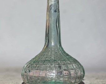 May include: A clear, light green glass vase with a bulbous base and a long, slender neck. The base features a raised, grid-like texture that creates a square pattern across the surface, while the neck remains smooth and cylindrical in shape.