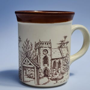 May include: Cream-colored ceramic mug with a brown rim and handle. The mug features a brown illustration of a church with a clock tower, trees, and a gate. A classic design for coffee or tea.