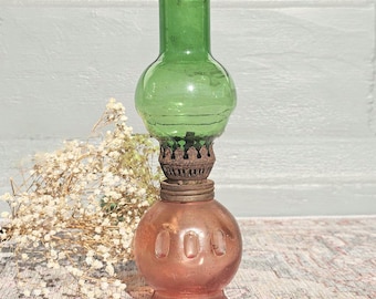 May include: An antique oil lamp with a green glass top and a reddish-orange glass base. The lamp has a metal connector and a decorative design. The lamp is a vintage item.