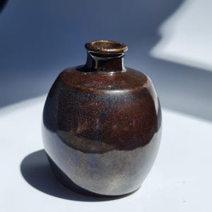 Puede incluir: Un pequeño jarrón de cerámica hecho a mano con un acabado marrón oscuro y brillante. El jarrón tiene un cuerpo redondeado y un cuello estrecho, perfecto para exhibir flores pequeñas o como elemento decorativo. El jarrón mide unos 10 cm de alto.