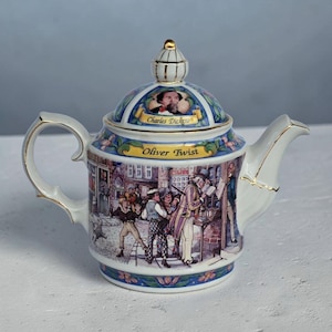 May include: A porcelain teapot decorated with scenes from "Oliver Twist." The lid features a portrait of Charles Dickens. The teapot has a white body with gold accents and a handle. The design includes a street scene with figures and the text "Oliver Twist."