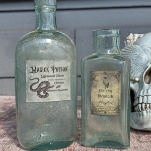 May include: Two clear, light green glass bottles with labels. The larger bottle features a snake graphic and text reading Magick Potion Medicinal Venom for the treatment of Chilblains AND Attitudinal Dysfunction. The smaller bottle reads Poison Hemlock.