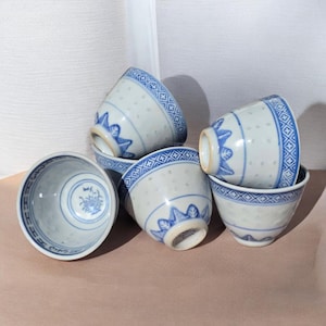 May include: A set of five white ceramic tea cups featuring blue hand-painted patterns. Each cup displays a geometric border at the rim, a series of small translucent dots, and a stylised leaf motif near the base. One cup shows a floral design inside.