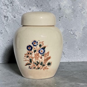 May include: A cream-colored ceramic jar with a matching lid, featuring a crackled glaze finish. The front of the jar is decorated with a detailed botanical illustration of blue flowers and brown foliage, centered on the rounded body of the vessel.