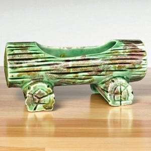 May include: A ceramic log-shaped planter in shades of green and brown. The planter has a hollowed-out center and is supported by two pairs of stylized feet. The surface is textured to resemble wood grain, adding a rustic touch to the decor.