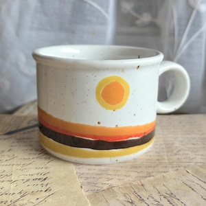 Op de afbeelding: Een witte keramische mok met een retro design. De mok heeft een gele zon en horizontale banden in oranje, bruin en geel. De mok heeft een oor en is bespikkeld met kleine bruine stippen.
