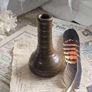 May include: A dark brown ceramic vase with a narrow neck and a wide, rounded base. The vase is sitting on a table with a piece of paper and a feather pen.