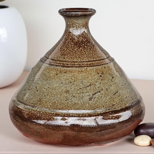 May include: A handmade ceramic vase with a distinctive form and an earthy colour scheme. The vase has a dark brown base and rim, leading to a lighter green-brown body. Horizontal lines encircle the vase as a decorative element.