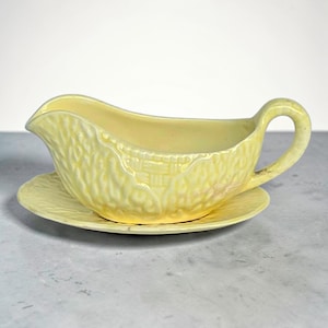 May include: A pale yellow ceramic gravy boat with a matching saucer. The gravy boat has a textured exterior and a curved handle. The saucer is round and also textured. The set is designed for serving sauces and gravies.
