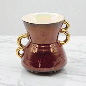 May include: A decorative vase with a metallic bronze finish and gold handles. The vase has a wide, flared top and a rounded base. The interior is a pearlescent white. The handles are circular and add a touch of elegance to the design.