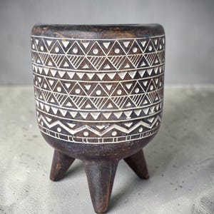 May include: A dark brown wooden planter with three short legs and a cylindrical body. The exterior surface features carved white geometric patterns, including rows of triangles, dots, and horizontal lines encircling the entire vessel.