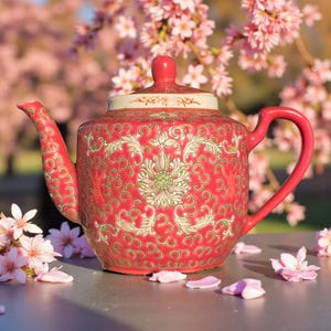 May include: A red ceramic teapot with a floral design and gold detailing. The teapot has a matching lid and handle. Pink flower petals are scattered around the base. The background features pink blossom trees.