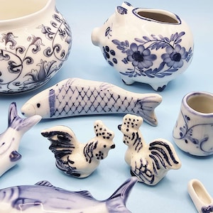 Może przedstawiać: Kolekcja białych ceramicznych figurek z niebieskimi wzorami kwiatowymi i rybami. Asortyment obejmuje dzbanek, skarbonkę, ryby, koguty i małe buty. Elementy są ułożone na jasnoniebieskiej powierzchni.