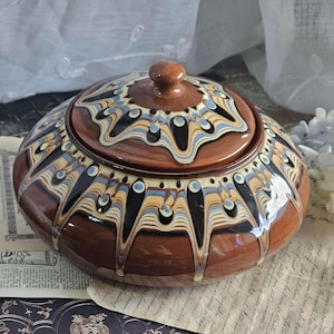 Puede incluir: Una jarra de cerámica marrón con tapa, decorada con un patrón de espiral de negro, blanco, azul y amarillo. La jarra tiene un diseño tradicional de arte popular.