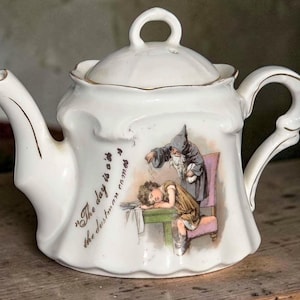 May include: A white porcelain teapot with gold trim, featuring a lid with a loop handle. The body displays a colourful illustration of a figure in a grey cloak standing over a seated child, with the text "The day is o'er, the dustman comes."