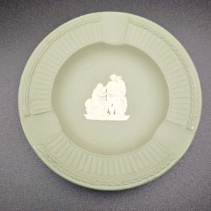 Può includere: Un posacenere in ceramica verde con un designo a rilievo bianco di tre figure. Il posacenere ha un bordo rialzato con un motivo decorativo.