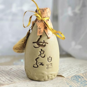 Puede incluir: Una pequeña botella de cerámica blanquecina con caracteres chinos negros y una cinta dorada atada alrededor del cuello. La botella tiene una superficie texturizada y una pequeña etiqueta. Está sobre papel vintage.