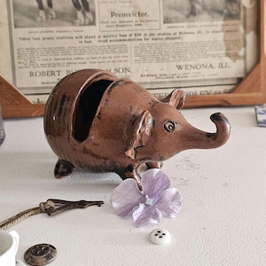 May include: A brown ceramic elephant figurine with a textured surface and a small hole in the top. The elephant is standing on a white surface with a purple flower and a white button.