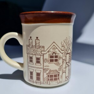 May include: Cream-colored ceramic mug with a brown rim and handle. The mug features a detailed brown illustration of houses and buildings. The mug is designed for hot beverages and is suitable for home or office use.