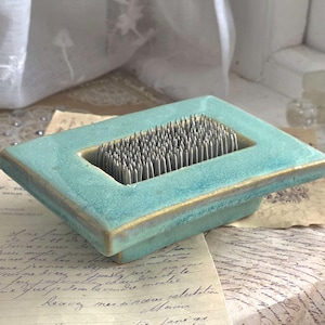 Może przedstawiać: Turkusowy ceramiczny żabnik do kwiatów z metalowymi szpilkami. Prostokątny żabnik ma zagłębiony obszar wypełniony szpilkami do układania kwiatów. Krawędzie są ciemniejsze turkusowe ze złotymi akcentami.