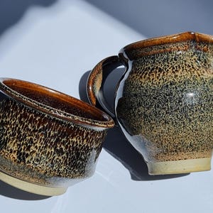 May include: A handmade ceramic creamer and bowl set. The creamer has a handle and spout, and the bowl has a wide rim. Both pieces are glazed in a speckled brown and tan pattern, with a light beige base. The set is ideal for serving coffee or tea.
