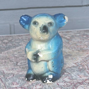 May include: A vintage blue and cream-colored koala bear figurine. The koala has a light cream-colored face and chest, with blue ears and body. The figurine has black accents on the nose and feet, with some paint chipping.