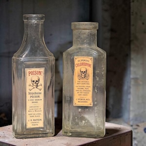 May include: Two clear glass apothecary bottles with rectangular bodies and narrow necks. The left bottle features a label reading POISON! Strychnine POISON. The right bottle features a label reading DR. SPENCER'S CHLORODYNE with a skull and crossbones.
