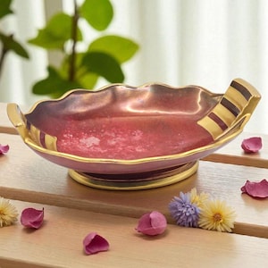 May include: A decorative, scalloped-edge bowl with a pink interior and gold trim. The bowl has gold handles with brown stripes. The bowl is on a wooden surface with flower petals and small flowers.