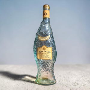 May include: A clear glass bottle shaped like a fish with a textured scale pattern. The bottle features a gold-coloured cap and two labels with the text Pescevino Salmon Blush, Vendemmia, and Product of Italy, with a volume of 750 ml.