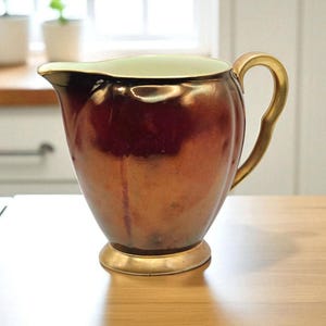 May include: A vintage-style pitcher with a bronze-colored exterior and a gold-colored handle and base. The interior has a light green color. The pitcher is likely made of ceramic or porcelain and is suitable for serving beverages.