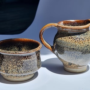 May include: A ceramic creamer pitcher and a small bowl. The pitcher has a handle and spout, and the bowl is round. Both pieces are glazed in a speckled brown, tan, and black pattern, with a lighter base.