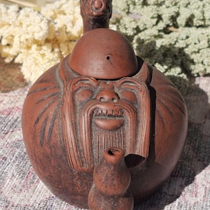 Puede incluir: Tetera de cerámica marrón con un diseño de rostro sonriente, con una larga barba y una cabeza redondeada. La tetera tiene un pequeño pico y un asa con un rostro detallado. El diseño general recuerda a una deidad asiática.