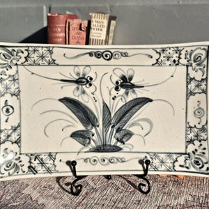 Puede incluir: Plato rectangular de cerámica con fondo blanco y diseño floral azul. El plato tiene un borde decorativo y está sostenido por un soporte de metal negro. El diseño floral presenta hojas y flores estilizadas.