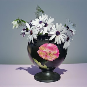 Puede incluir: Un jarrón negro con un diseño floral rosa y dorado contiene flores blancas con centros morados. El jarrón está sobre una superficie de color lila claro, con una sombra detrás.