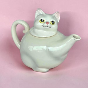 May include: A white ceramic teapot shaped like a seated cat. The lid forms the cat's head with yellow eyes, a pink nose, and a small smile. The body of the teapot is round with a curved handle on the left and a spout extending from the right side.