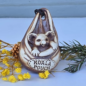 May include: A ceramic koala pouch figurine with a brown and cream color scheme. The koala is in a pouch with the words "KOALA POUCH" on the front. The figurine is decorated with yellow flowers and green leaves.