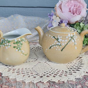 May include: A cream-coloured ceramic teapot and creamer set. The teapot has a lid and a curved handle. Both pieces are decorated with green and white floral designs. The set is placed on a white lace doily.