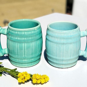 May include: Two ceramic mugs in a barrel shape, one teal and one light blue, with matching handles. The mugs are on a white surface, with yellow flowers and green stems in the foreground.