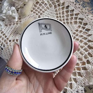 Puede incluir: Un pequeño plato de cerámica blanca con un borde negro y el texto "M.M. LINE" debajo de un emblema de bandera. El plato se sostiene en una mano con un anillo enjoyado azul y verde. El fondo presenta un tapete de encaje.
